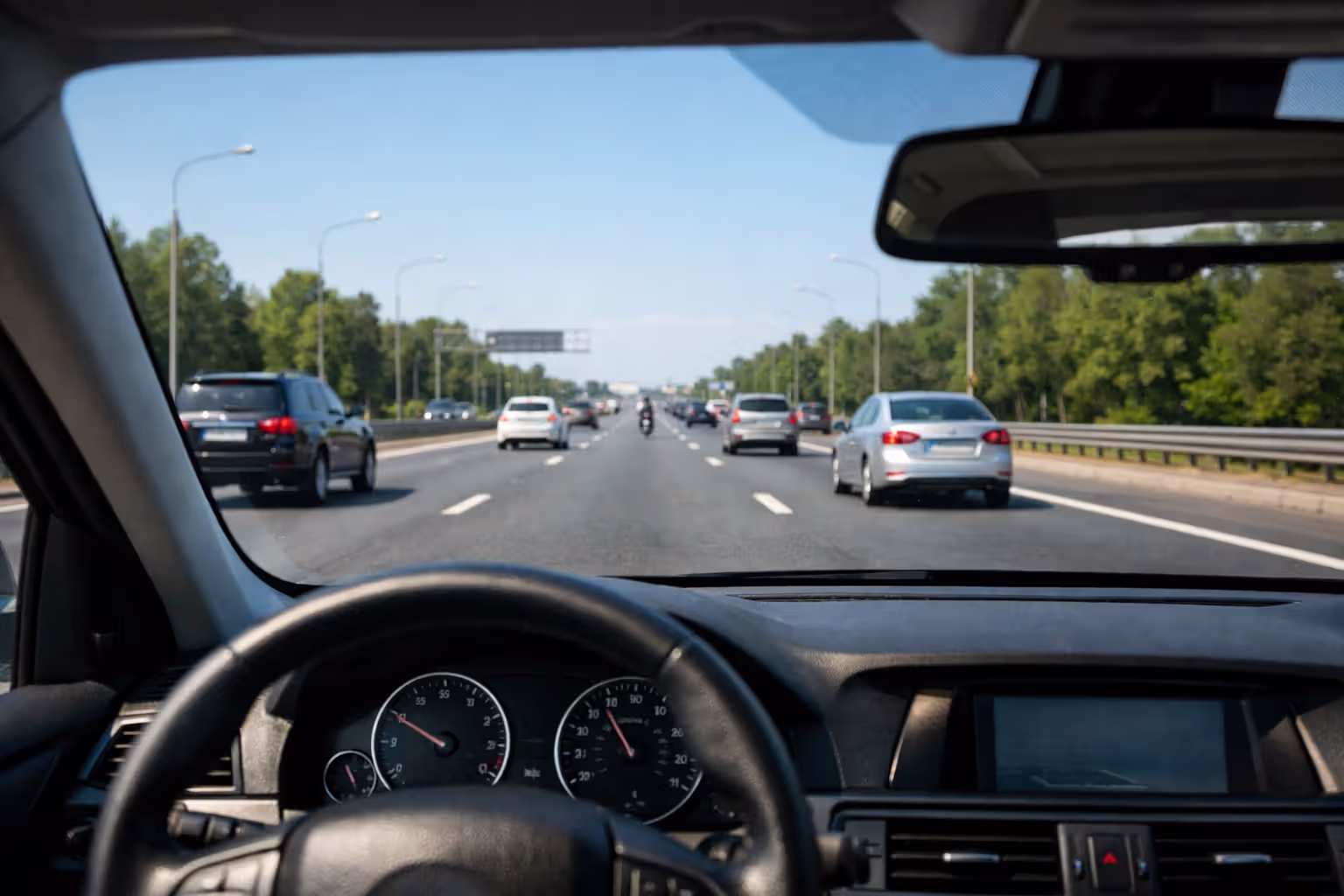 View from inside a car through windshield showing a distant approaching motorcyclist barely visible among traffic on a multi-lane road demonstrating inattentional blindness